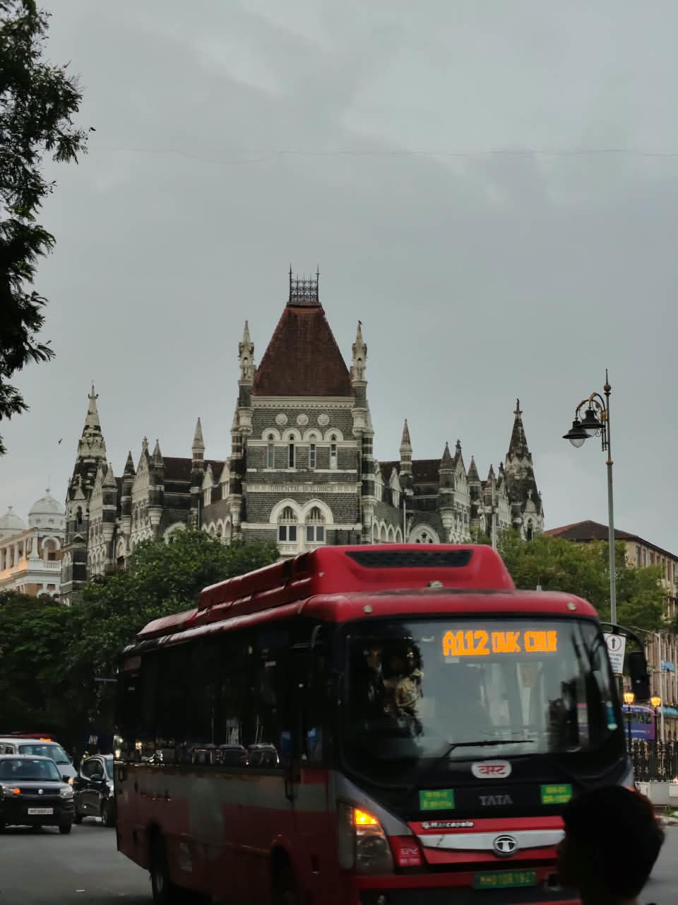 A red city bus passing the Oriental Buildings in Mumbai under a grey monsoon sky